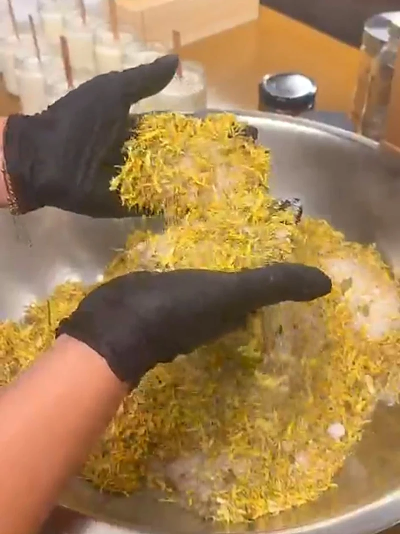 Gloved hands blending mineral salts and dried yellow botanicals in a large metal bowl on the studio workbench.