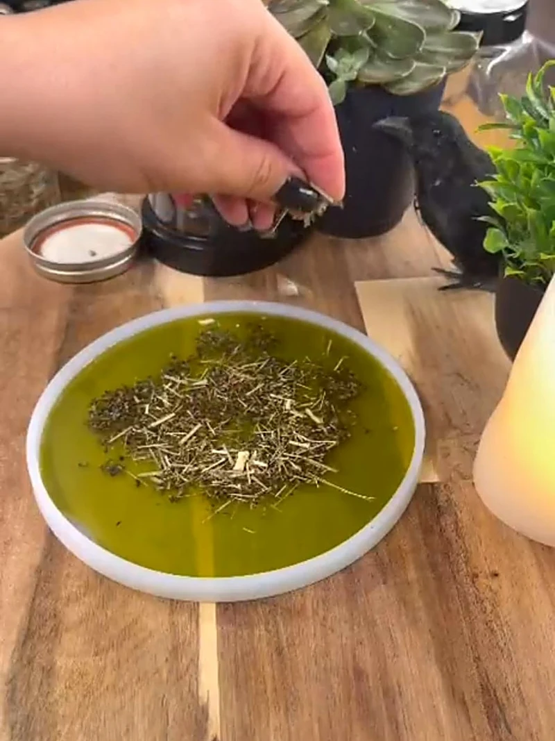 A hand setting dried herbs into a shallow bowl of warm green wax on the wooden studio workbench.