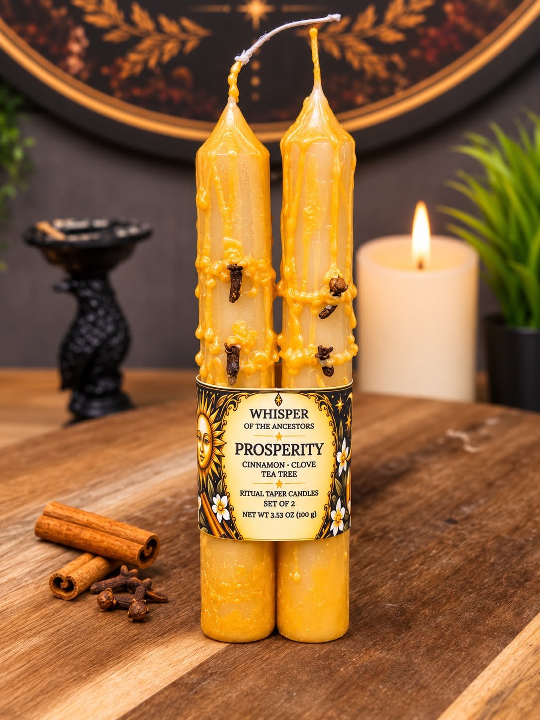 Pair of golden Prosperity ritual taper candles with heavy textured wax drips and whole cloves pressed into the wax, held together by a parchment and black label band, with rolled cinnamon sticks and loose cloves beside them